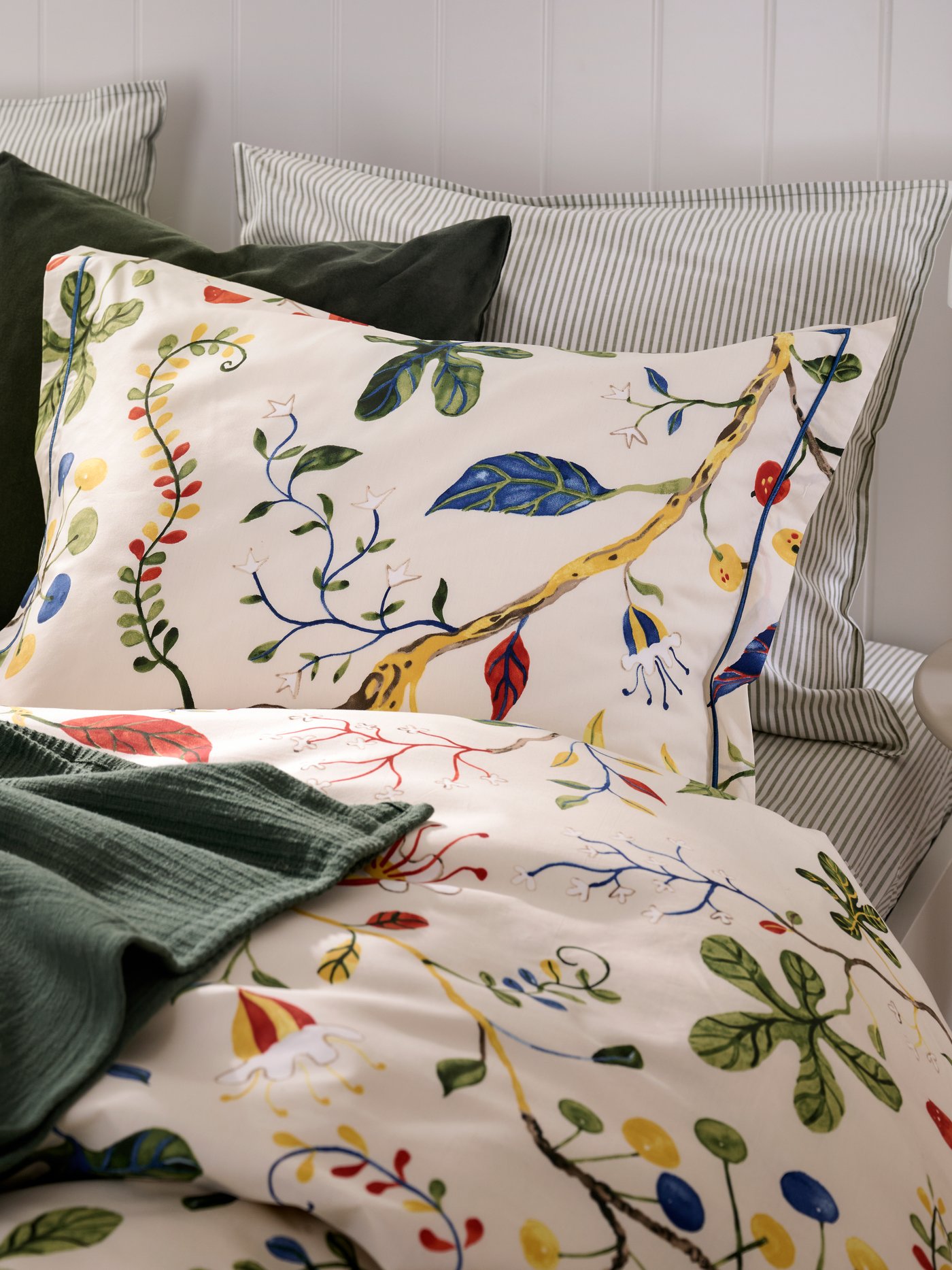 Close up of a bed featuring KRONBUSKE duvet and pillow cover and a green blanket, and a small bedside table.