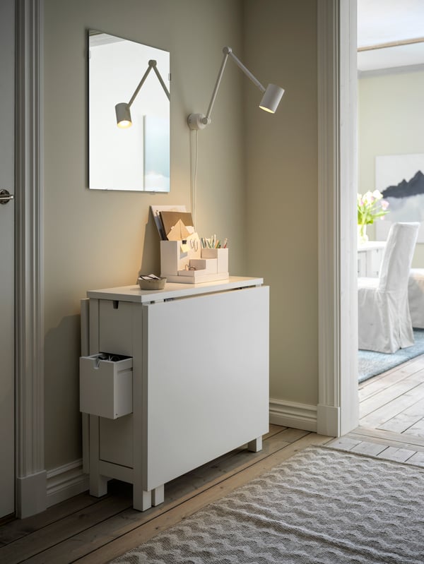 Un pasillo con una mesa NORDEN blanca plegable contra la pared y un espejo LÄRBRO con una lámpara NYMÅNE blanca encima.
