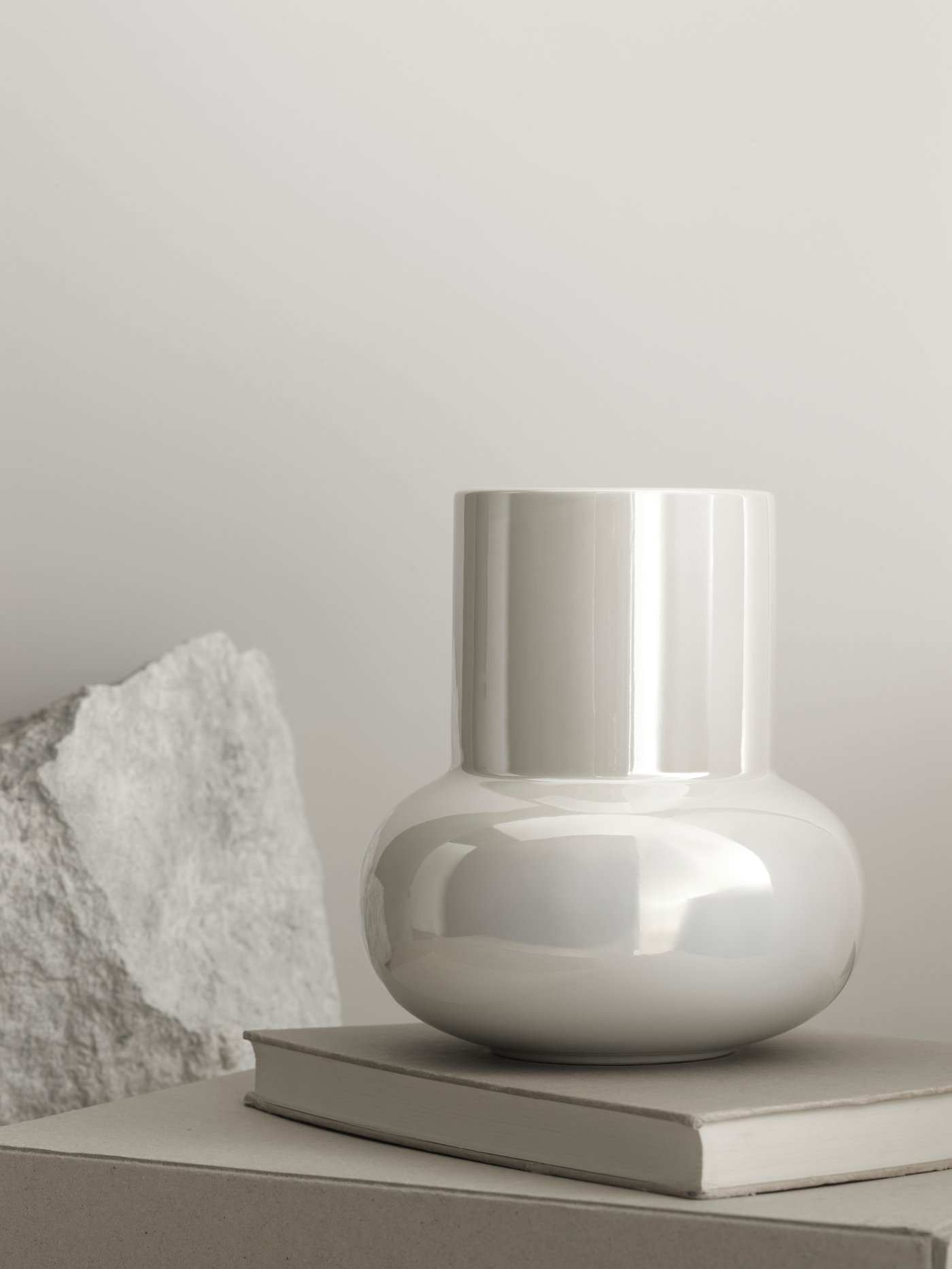The iridescent white FNITTRIG vase with a rounded shape placed on a surface with books and chunks of raw stone.