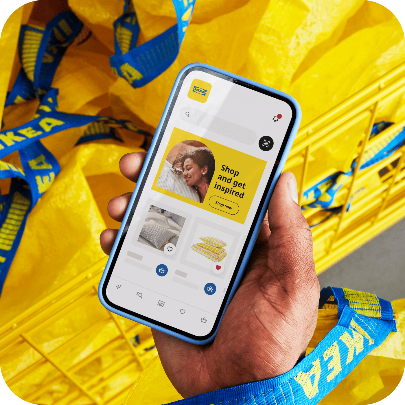Eine Person hält ein Smartphone in der Hand, auf dem die IKEA App geöffnet ist. Im Hintergrund sieht man einen Korb voll mit gelben FRAKTA Taschen.