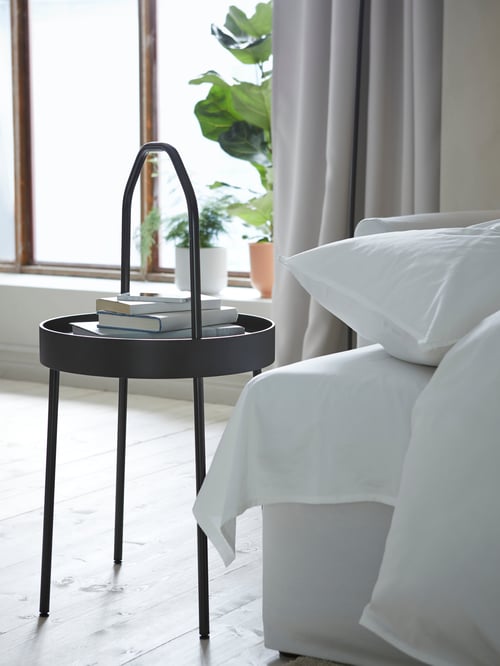 A black BURVIK circular side table with a thin, bent handle holding books and a cell phone by a sofa-bed with bedding.