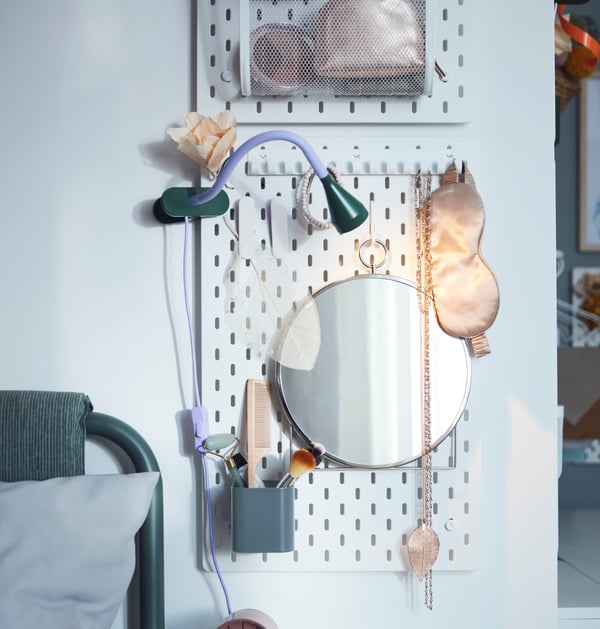 A circular mirror and a storage container hang on a  white SKÅDIS pegboard, holding beauty tools, jewellery and accessories.