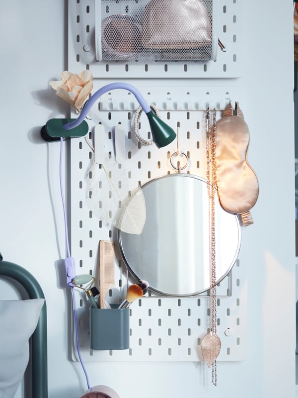 Un miroir circulaire et une boîte de rangement sont suspendus à une patère blanche SKÅDIS, où sont rangés des outils de beauté, des bijoux et des accessoires.