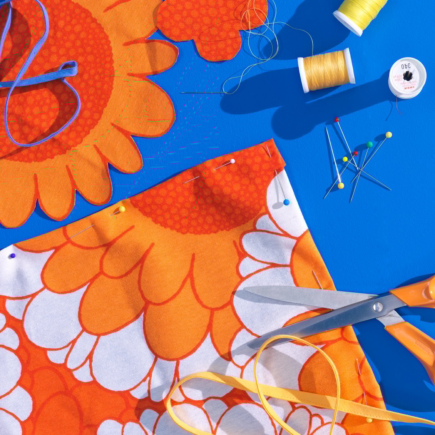 Fabrik prapotong 3 meter SANDETERNELL berwarna jingga terletak di atas meja berwarna biru dengan jarum peniti, gunting dan gelendong benang di sekitarnya.