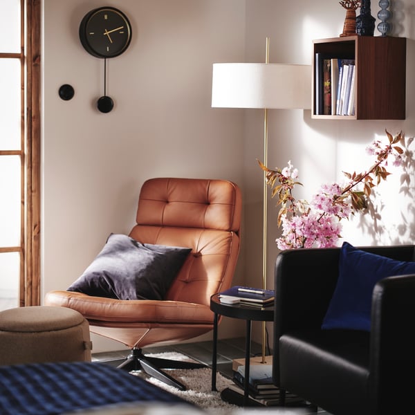 A corner of a bedroom with HAVBERG armchair in light brown, STOCKHOLM 2025 floor lamp and STURSK wall clock on the wall.