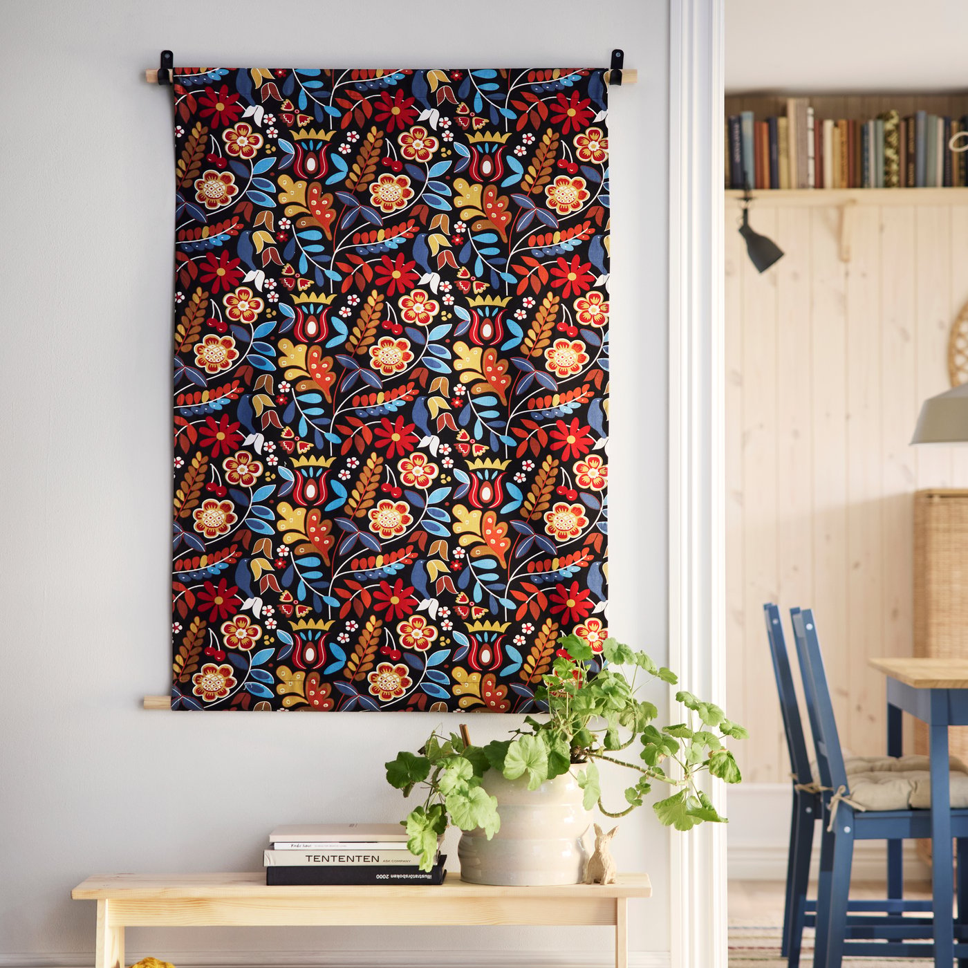 A wall decoration made from BLÅRAPUNKEL pre-cut fabric hangs on a wall above a plant in a beige HASSELBUKETT plant pot.