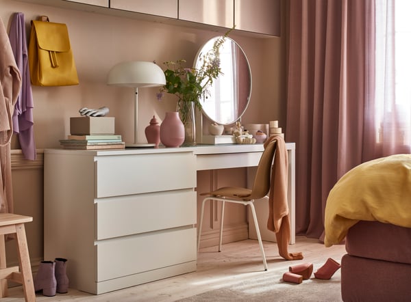 Sminkhörna med vitt skrivbord, rund spegel, beige stol med filt, och dekor i rosa och gröna toner mot en mjukt rosa väg