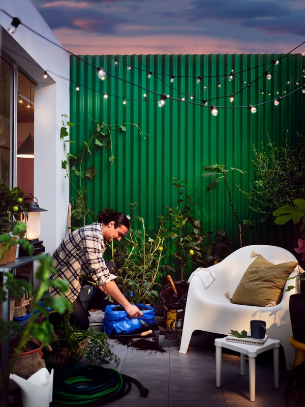 A patio dotted with plants, a white SKARPÖ armchair, an UTTER children’s stool, and a black UTSUND LED lighting chain.