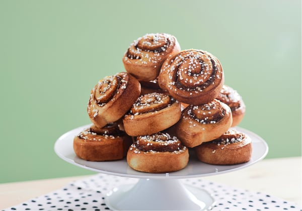 Pile de brioches à la cannelle fraîchement cuites, garnies de sucre perlé, présentées sur un plat blanc devant un fond vert clair.