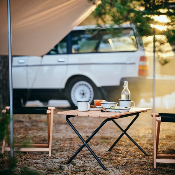 A SOLUPPGÅNG folding table with SOLUPPGÅNG plates and mugs on it is standing under a tarp, close to a car. 