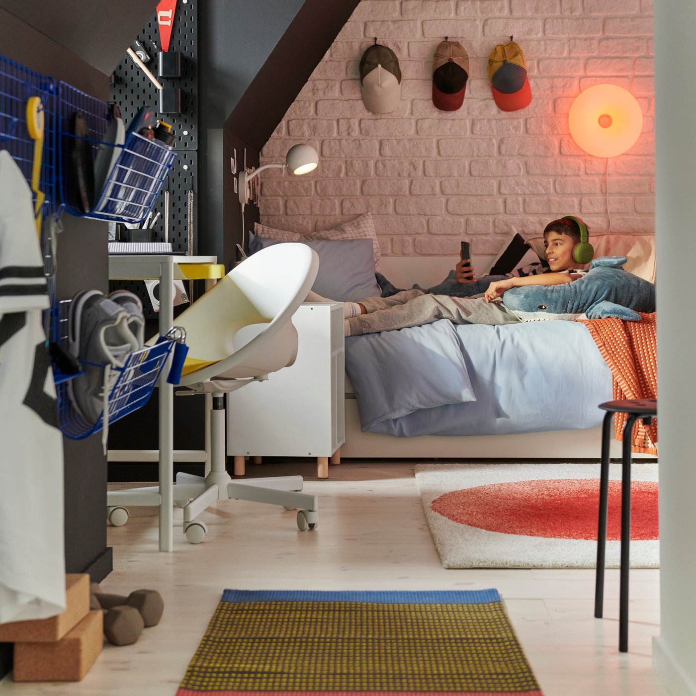A teenage boy lies on a VIHALS bed listening to music in headphones. In the foreground: a white ENKROKEN desk and a chair.
