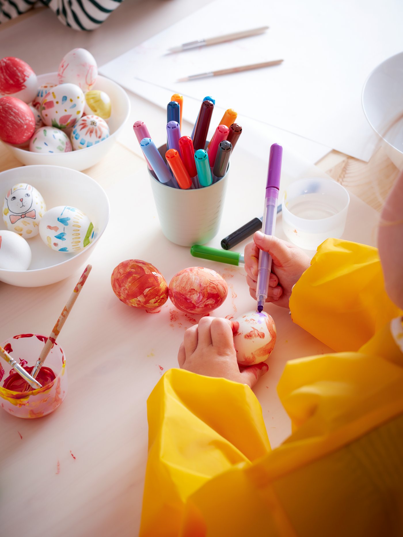Un niño vestido con un delantal amarillo de manga larga delante de un protector de escritorio blanco con rotuladores en una taza, pinceles y un cuenco.