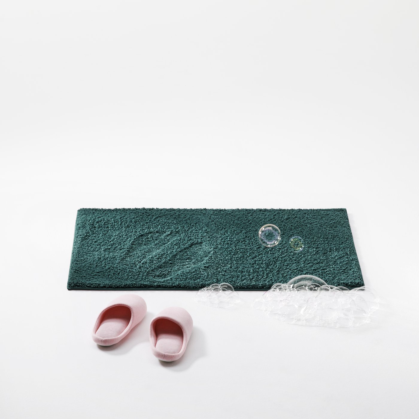 Rosa tøfler og såpebobler ved siden av en BACKMÅRA badematte i et rent, hvitt studiomiljø.