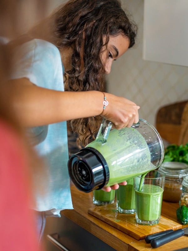 A young woman pours a green smoothie from a blender into two POKAL glasses. Her mother is standing next to her.