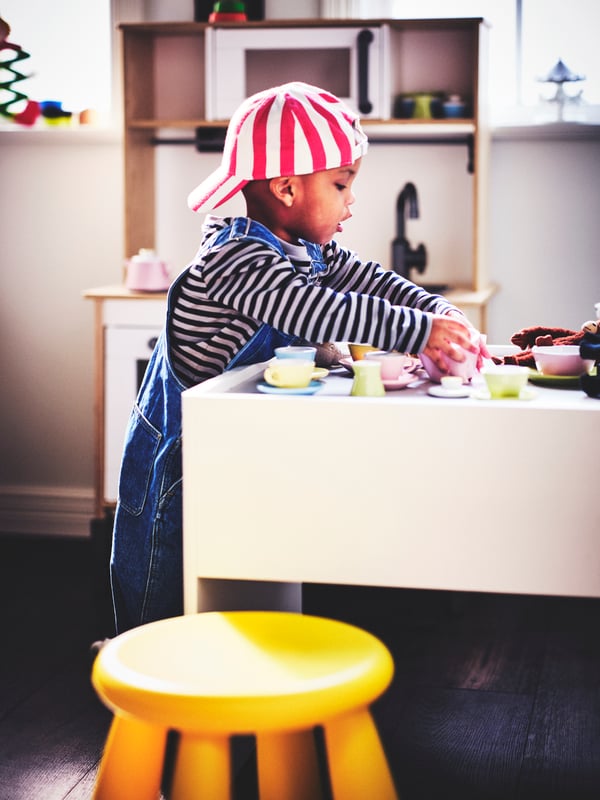 A child playing with a DUKTIG tea playset at a white DUNDRA activity table with a yellow MAMMUT children’s stool to the side.
