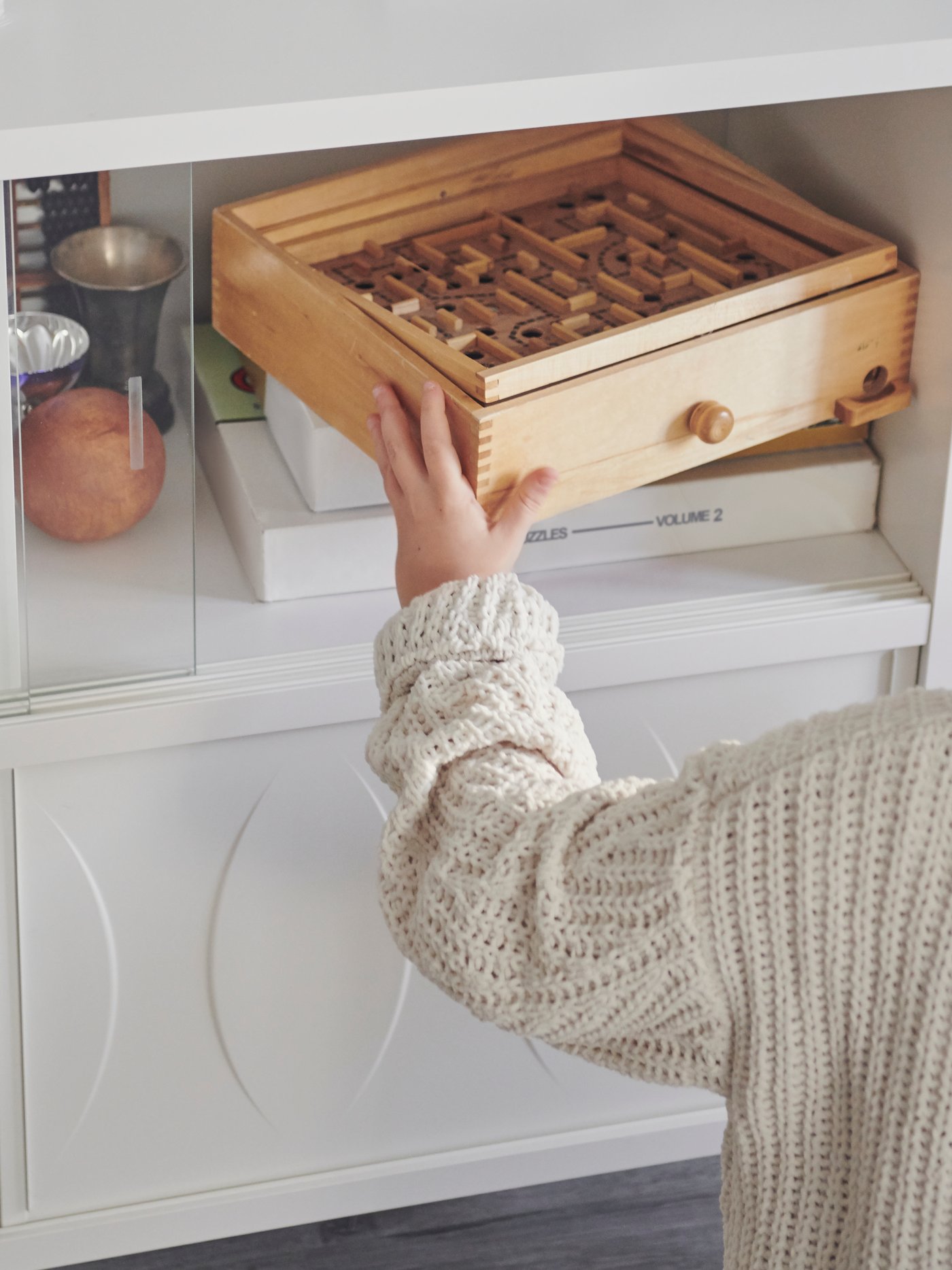 A child’s hand holds the corner of a wooden labyrinth game, halfway out of the top compartment of a white KALKNÄS cabinet.