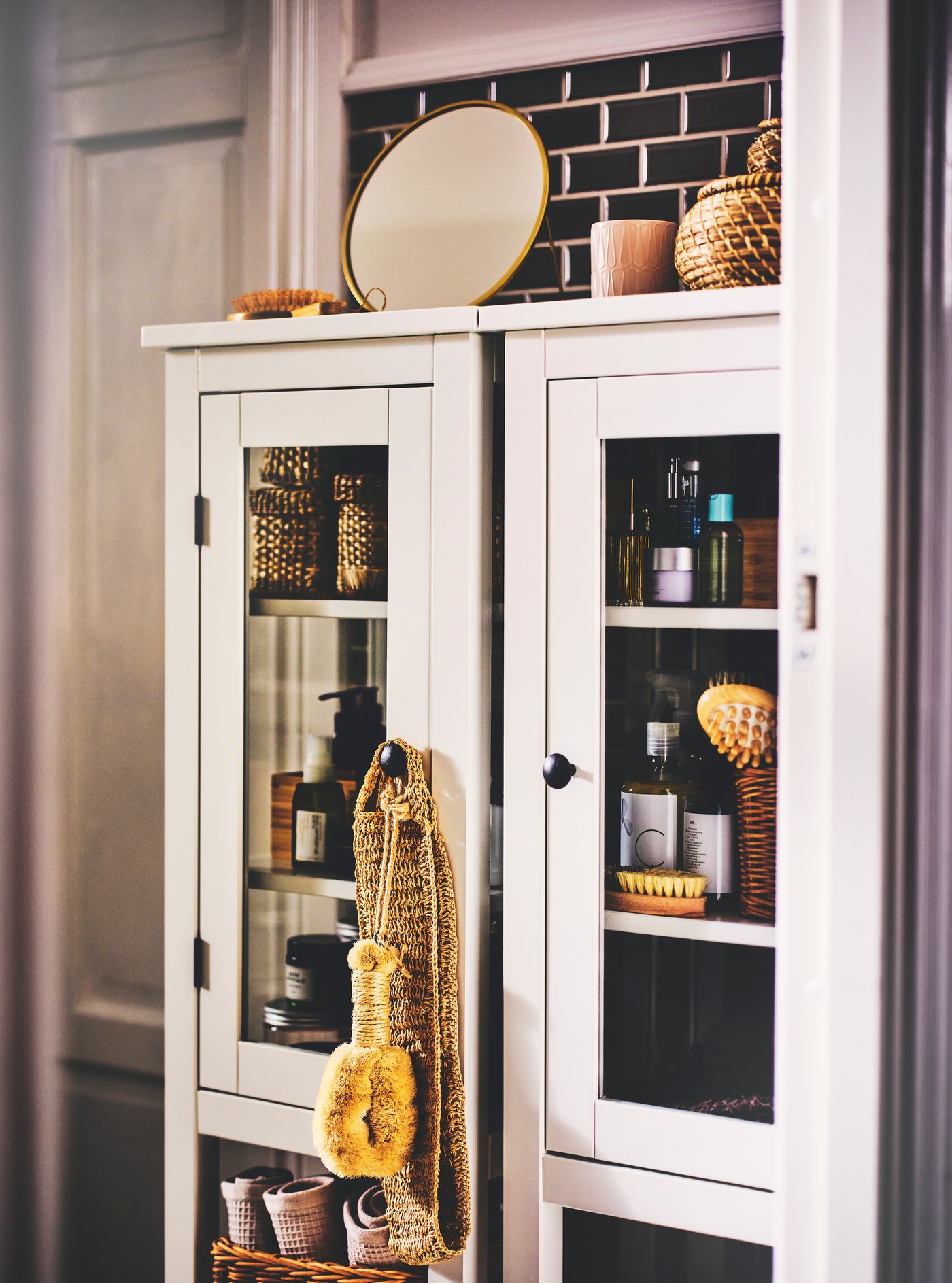 زوج من الخزائن المرتفعة بأبواب زجاجية HEMNES باللون الأبيض، وبداخلهما توجد مستلزمات حمام، وهناك صناديق بأغطية ومرآة في الأعلى.