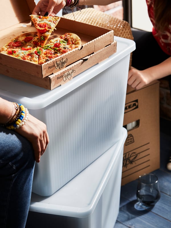 Two RYKTA storage boxes with lids are stacked on top of each other. People are sharing a pizza, placed ontop of the boxes.