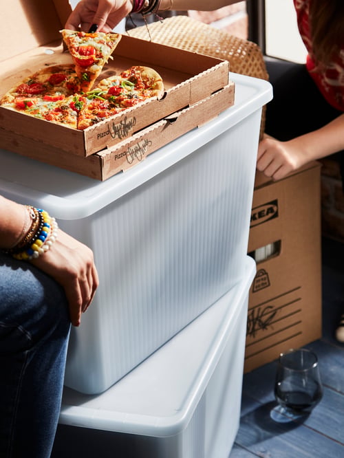 O bucătărie cu două persoane care mănâncă o pizza așezată pe o cutie transparentă de depozitare RYKTA gri-albastră cu capac.