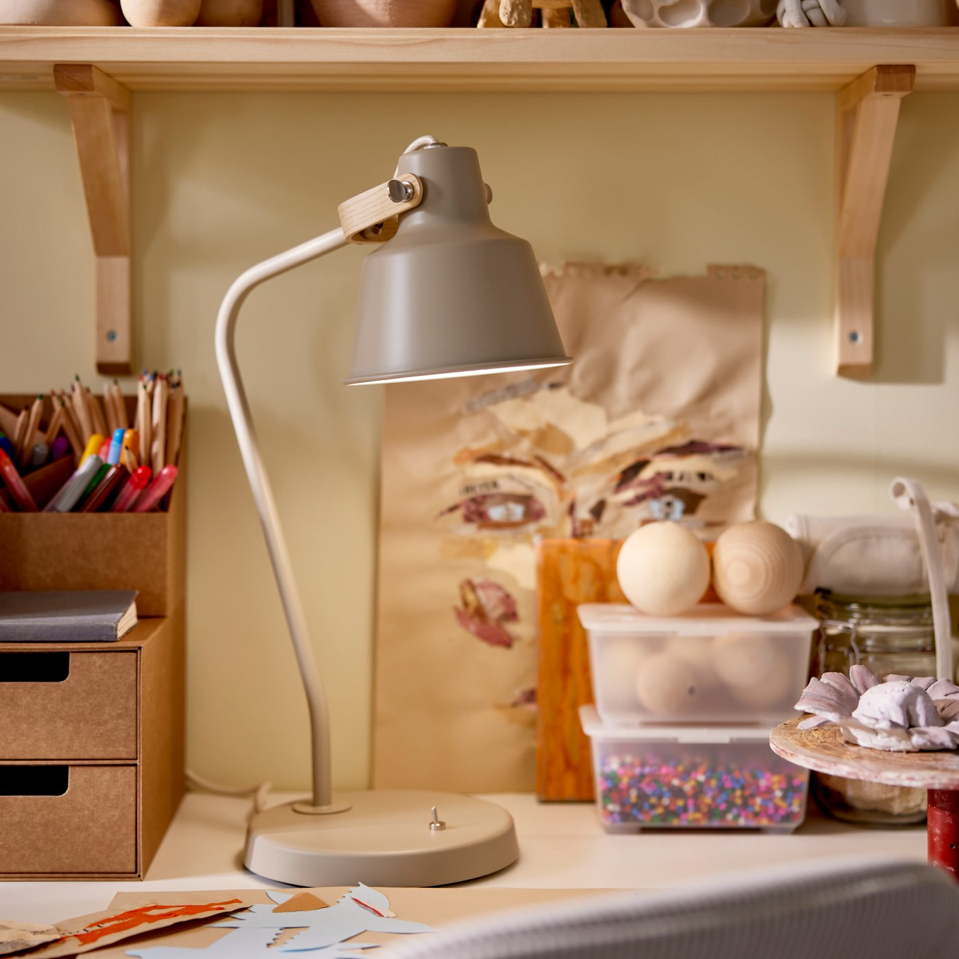 En tänd SJÖMAKT skrivbordslampa står på ett skrivbord med många föremål på under en TRANHULT/SANDSHULT vägghylla.