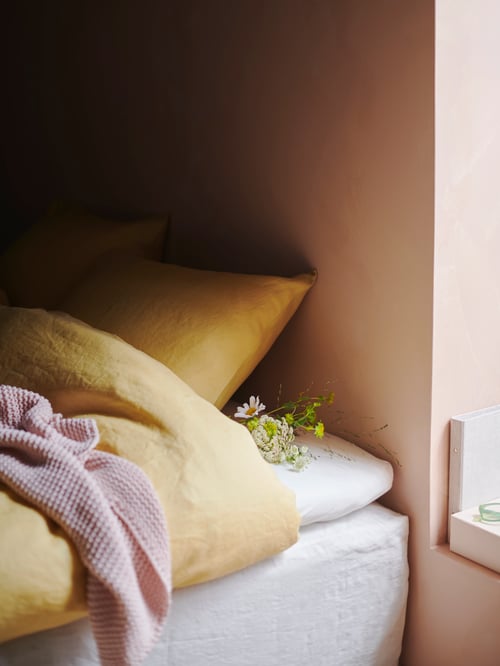 Chambre à coucher avec drap-housse blanc en coton placé sous une housse de couette jaune avec 2 taies d’oreiller et un jeté rose.