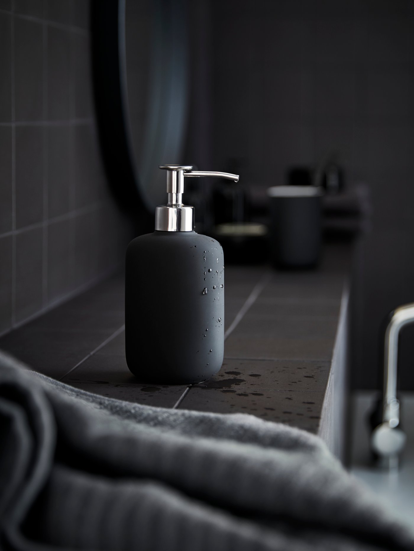 A dark grey EKOLN soap dispenser is standing in a grey bathroom, next to a mirror and with a grey towel in the foreground.