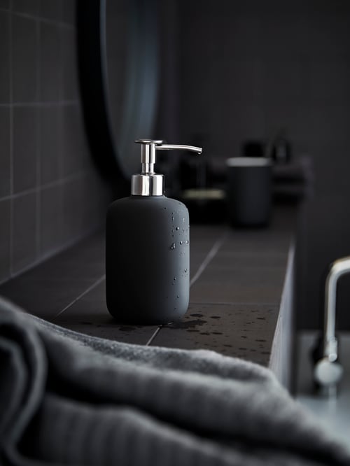 Espace salle de bains où se trouve un distributeur de savon gris posé sur une surface recouverte d’un carrelage gris. Il y a un textile gris à l’avant-plan. 