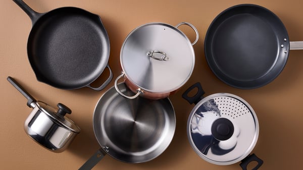 Pots and pans made of different materials, from cast iron to stainless steel, are placed on a light brown surface.
