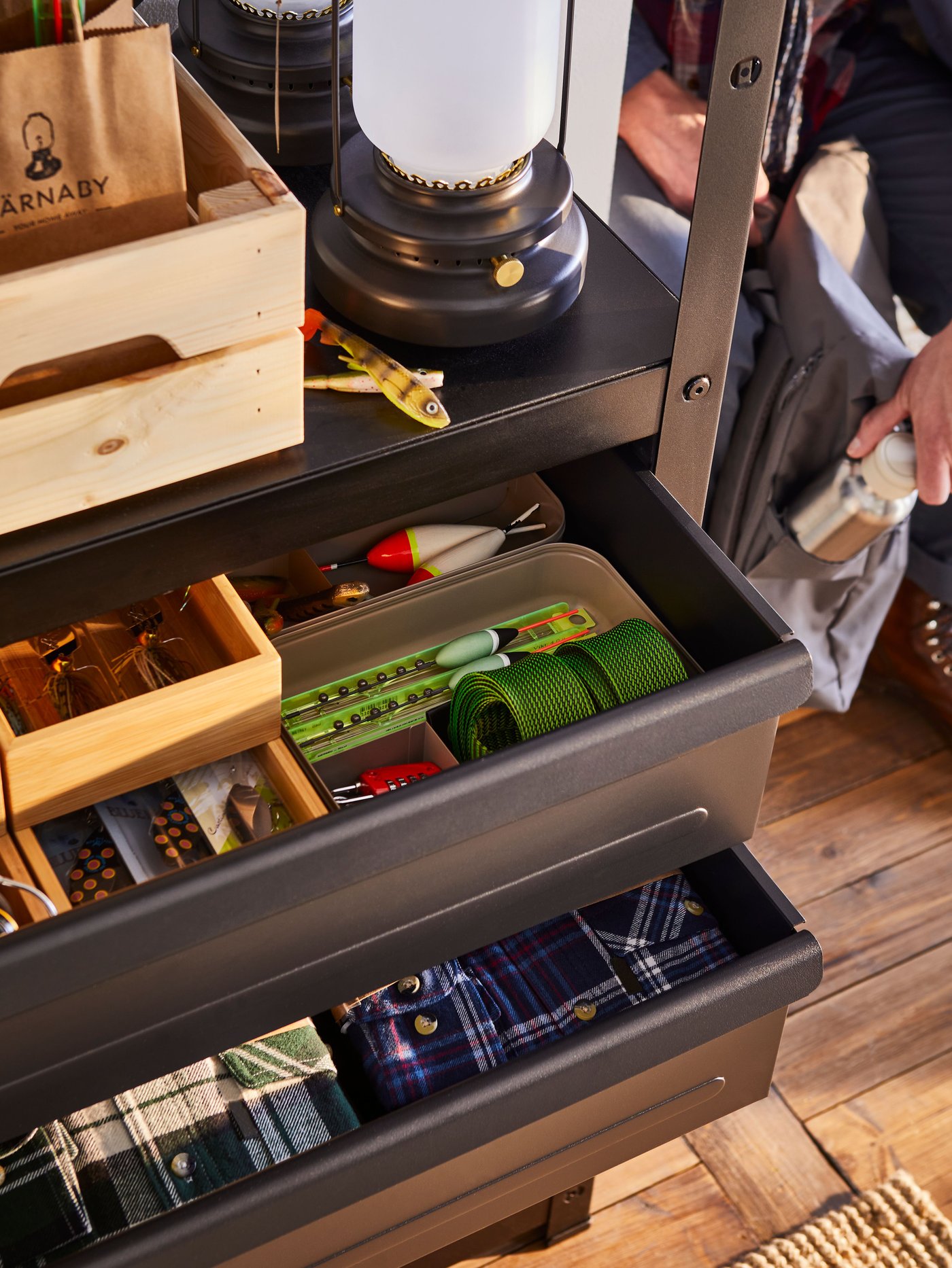 A BROR shelving unit with drawers/shelves stands with two open drawers containing folded shirts and fishing tackle.