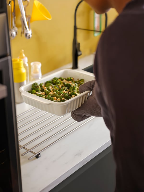 A hand with an oven glove puts an off-white oven dish on a LÄMPLIG stainless steel trivet on a white marble effect worktop.