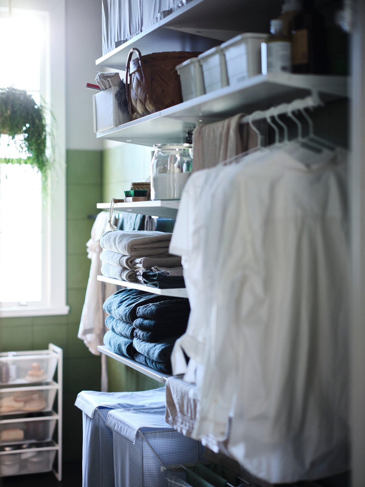 En närbild på en vit öppen BOAXEL garderobslösning med prydligt vikta handdukar på hyllor och vita skjortor som hänger i ett grönt badrum.