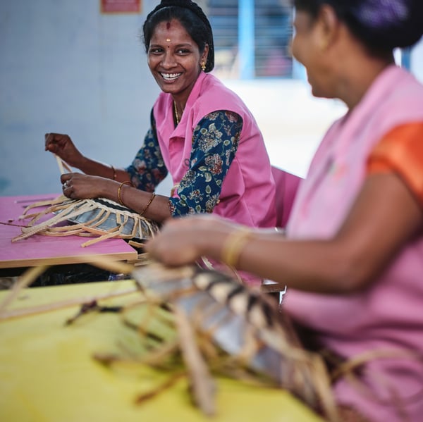 A person dressed in pink is hand braiding a MÄVINN wall decoration. She is smilingly looking at her friend.