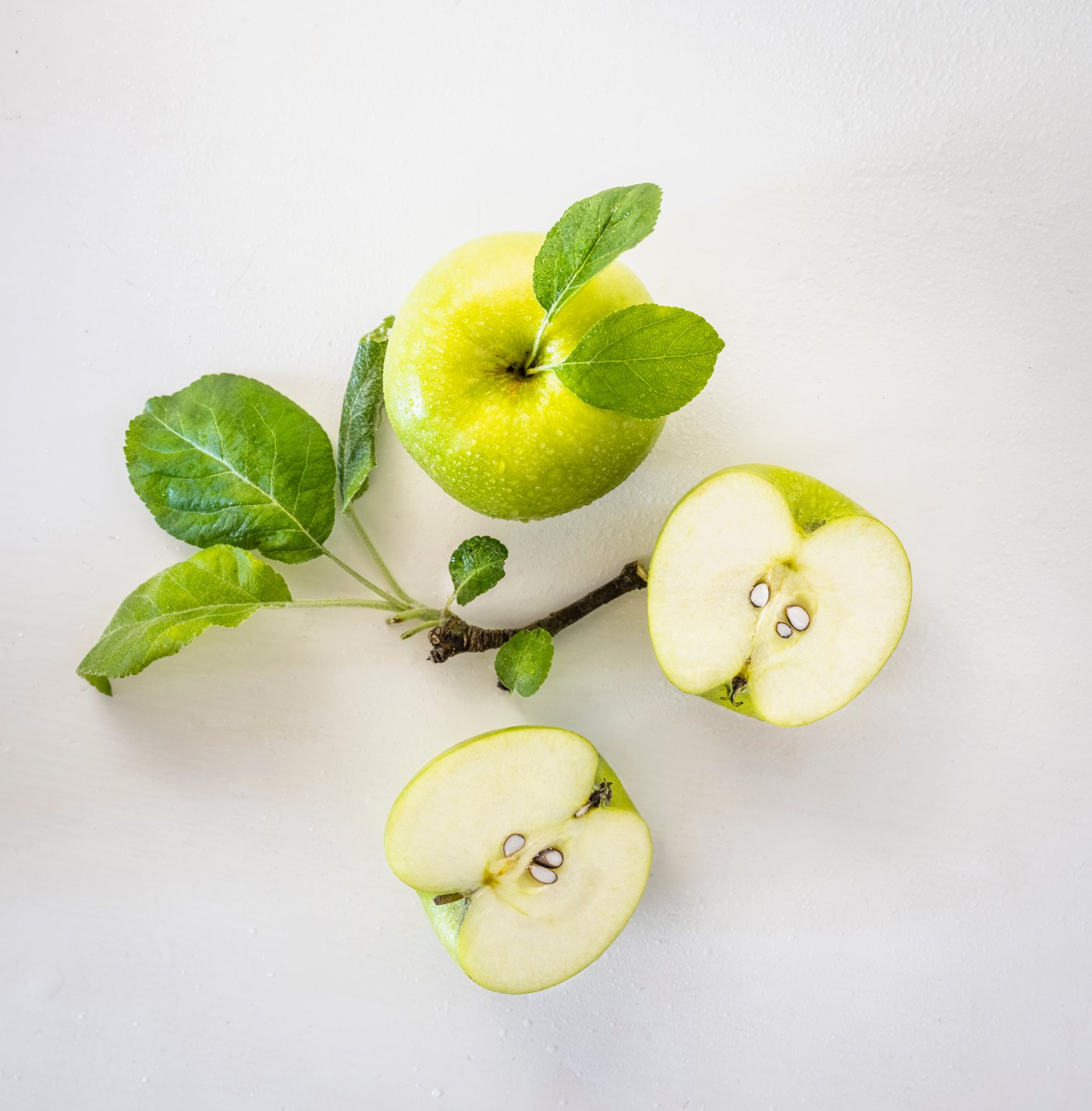 Mela verde intera con foglie e due metà di mela tagliata, disposte su fondo chiaro.