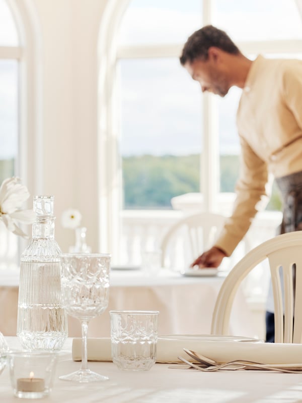 A man sets a table in a light and airy restaurant, by a table with a SÄLLSKAPLIG carafe with stopper, wine glass and glass.
