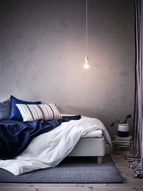 A SMALSTÄKRA cushion cover in beige and blue stripes, on a bed with some blue and white bedding and a hanging light-bulb.