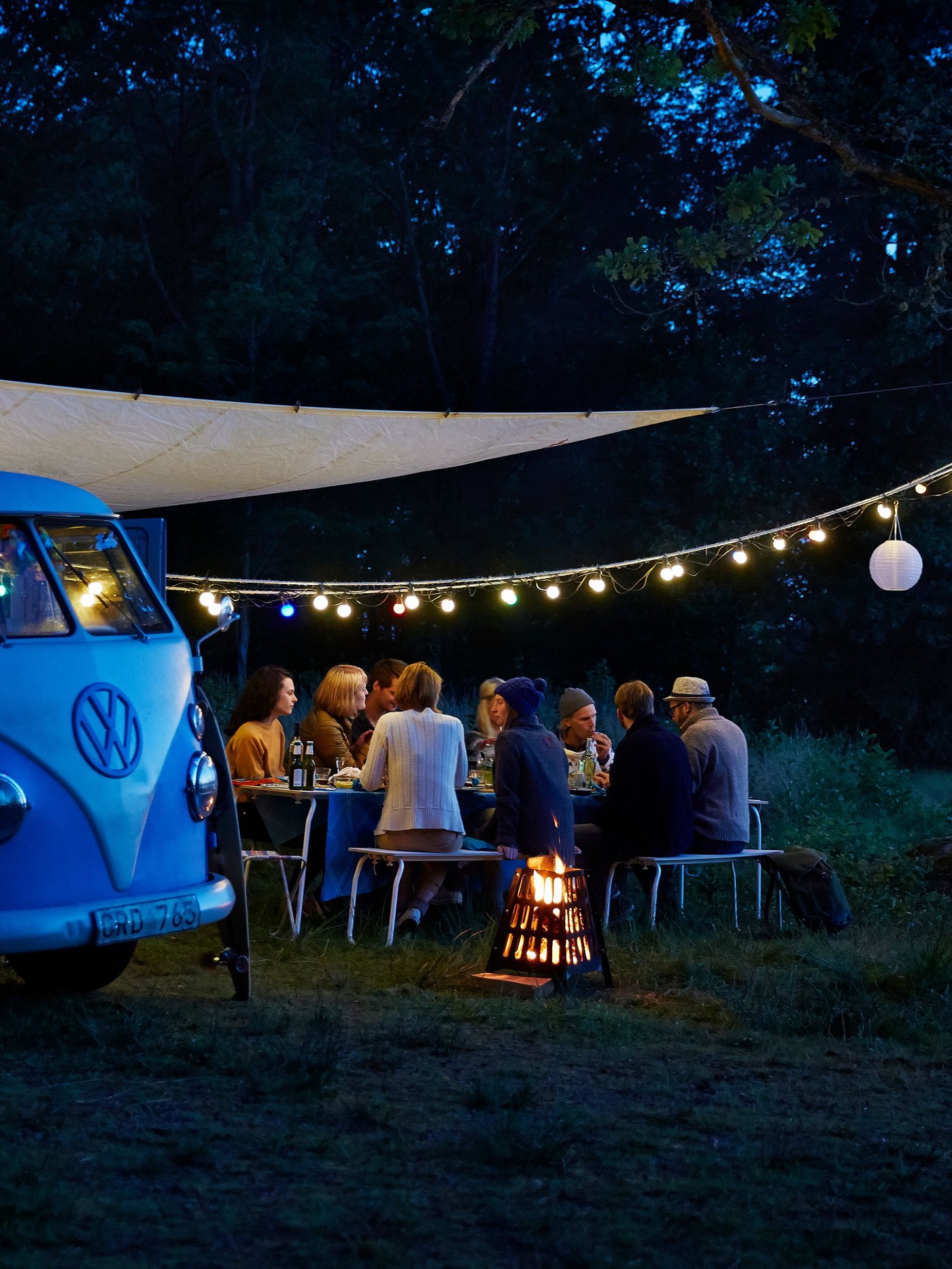 Eine Gruppe von Freunden geniesst ihr Abendessen im Freien neben einem alten blauen VW-Bus, der abends von Lichterketten und einem gemütlichen Feuer beleuchtet wird.