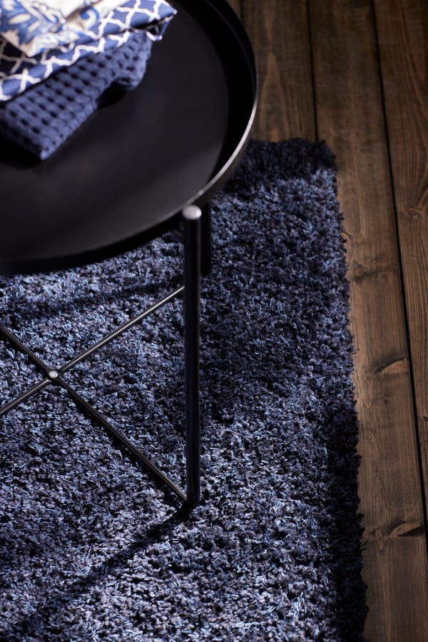 Zicht vanuit een donkerblauw fluwelen salon naar een venster met lange transparante witte gordijnen en donkere overgordijnen, en een glazen salontafel en plant.