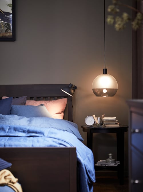 Un lit IDANÄS agrémenté d’une housse de couette et de taies d’oreiller PUDERVIVA bleu foncé est située près d’une table de chevet. Une suspension surplombe l’ensemble.