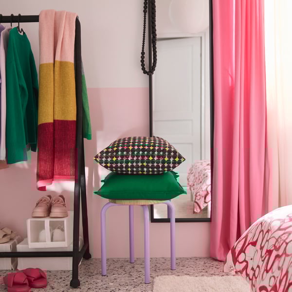 A bedroom with a pink/green ÄLVSJÖSKOGEN cushion cover placed on a green GURLI cushion cover stacked on a stool.