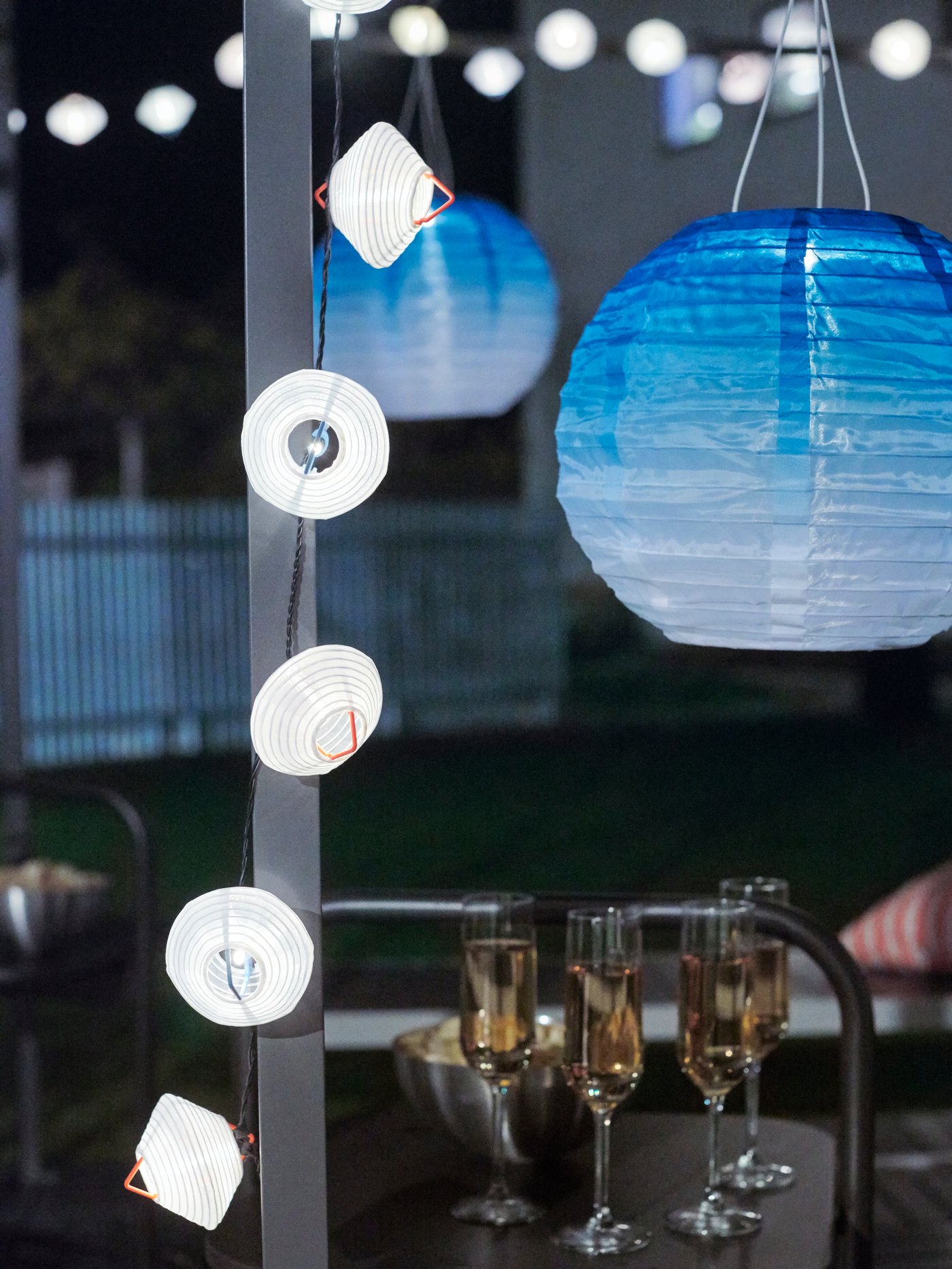 מנורות תלייה לד סולאריות SOLVINDEN בגוון כחול, שרשראות תאורה בצבע לבן, כוסות יין וחטיפים בגינה בשעת לילה.