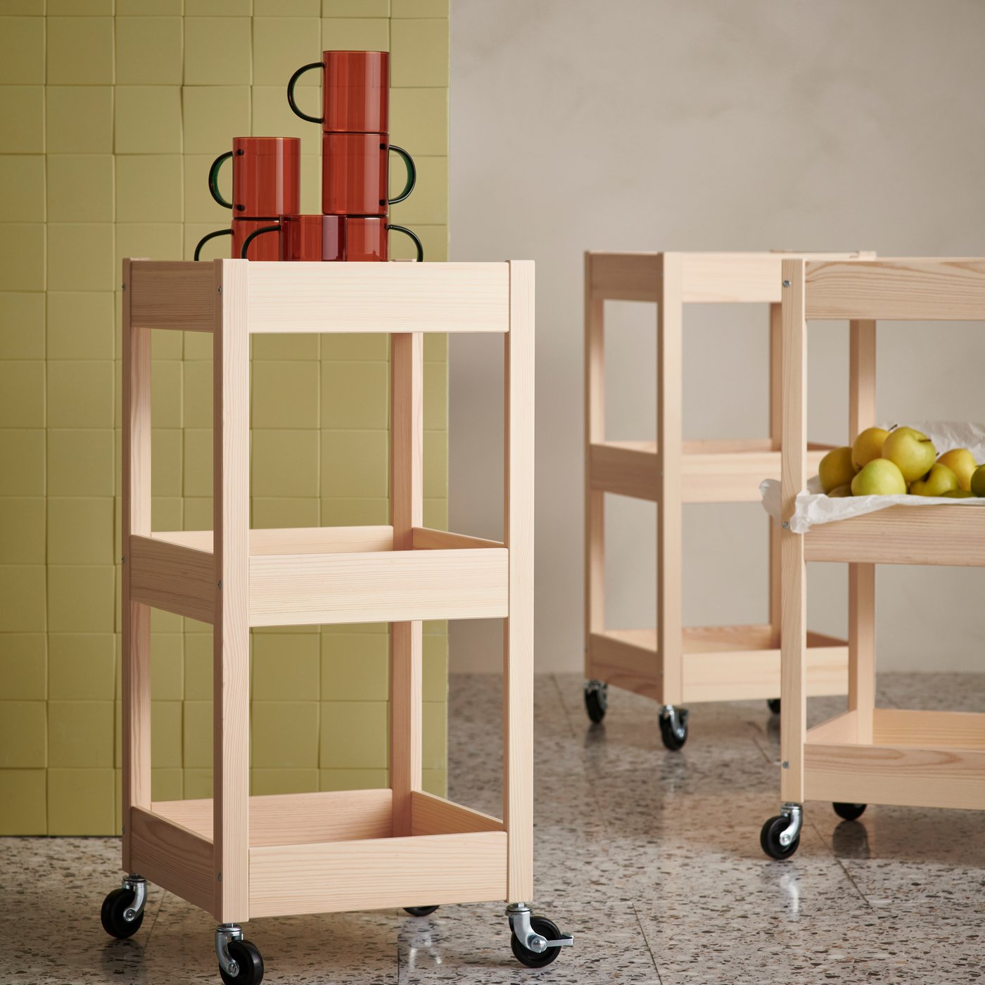 Three LOSHULT kitchen trolleys on castors. One with SKUGGSTUBB mugs on the top shelf, one with a bunch of apples. 