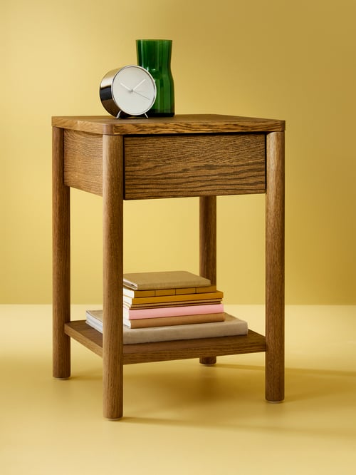 A green TIDVATTEN vase and a MALLHOPPA alarm clock are on a beige TONSTAD bedside table. A stack of books are underneath.