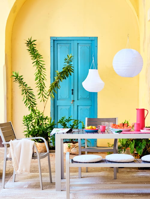 A terrace with a dining table laid for dinner, with FRÖSÖN/DUVHOLMEN cushions on the bench and lanterns above the table.