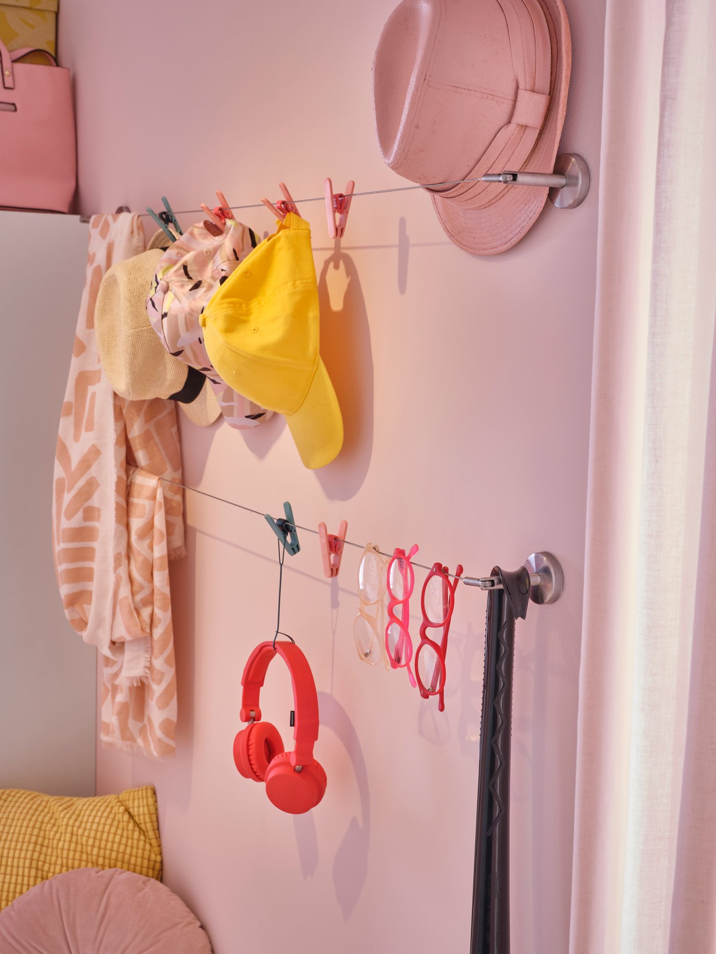 Dos cables de cortina sujetos a una pared rosa. Pinzas rosas y grises con gorros y bufandas amarillos y rosas, y unos auriculares rojos.