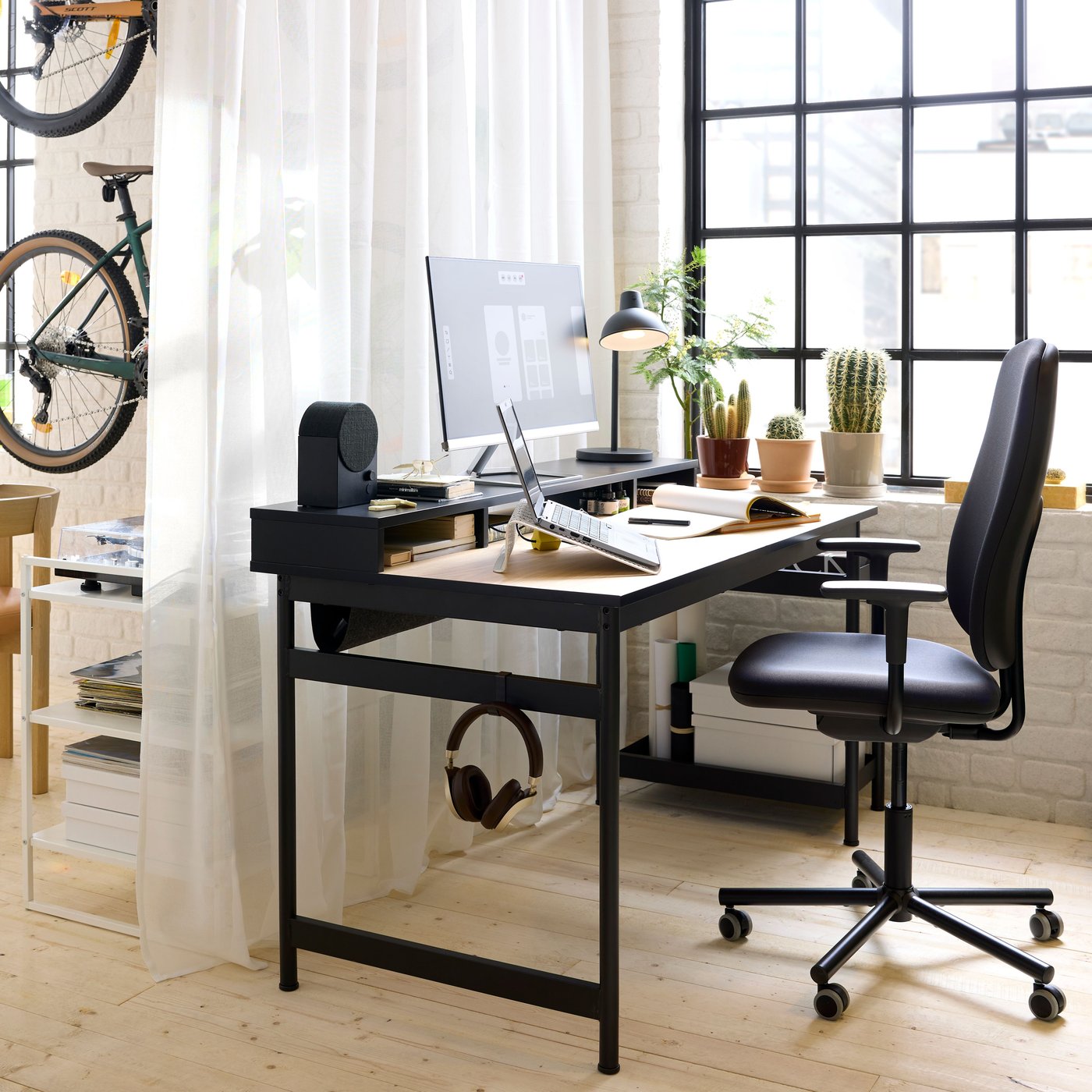 En mörkgrå SMÖRKULL kontorsstol med armstöd står vid ett MULLSJÖ skrivbord i ekfanér bredvid ett stort fönster.