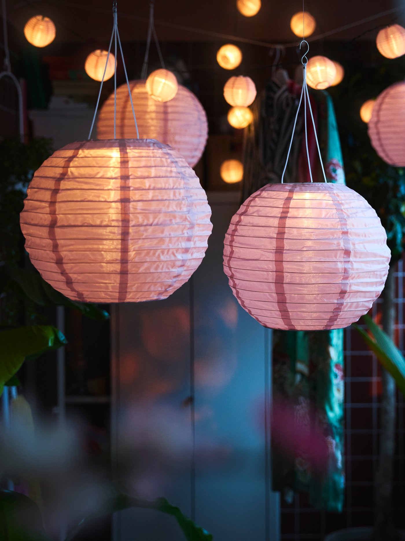 A balcony decorated with lush plants features several pink SOLVINDEN LED solar-powered pendant lamps hanging above.
