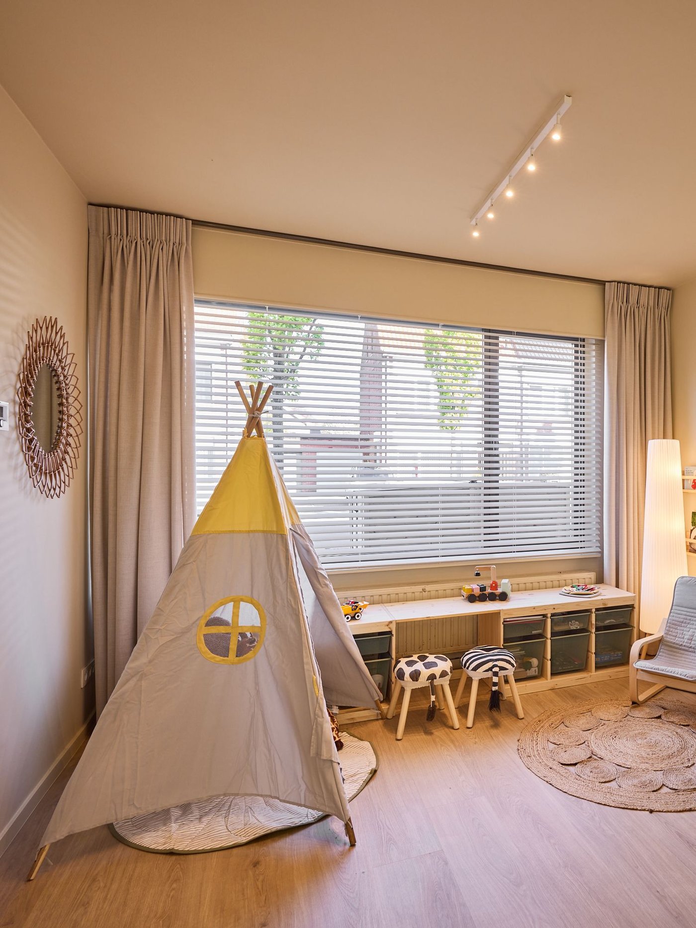 Speelhoek met tipi tent, lage opbergkast en krukjes bij raam met jaloezieën, in een gezellige en lichte kinderkamer.