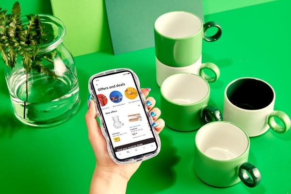 A hand with green-painted fingernails is holding a smartphone displaying the IKEA app over a green table with mugs.