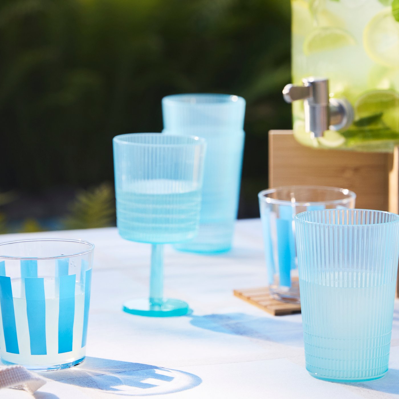 Blå KALLSINNIG og PILLESTARR glas på et havebord sammen med en IKEA 365+ kande fyldt med limonade.