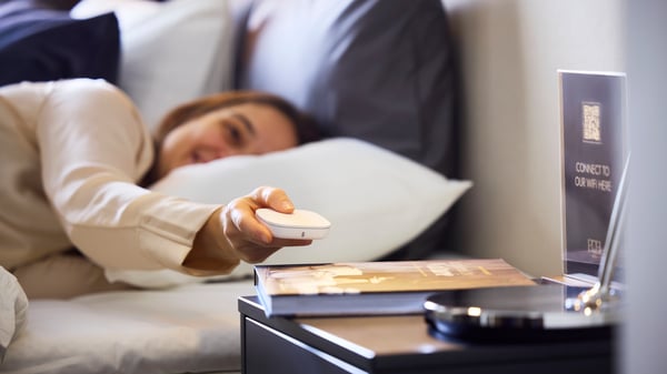 A person in a hotel bed holds a white STYRBAR remote, that is square with rounded corners, to control IKEA smart lighting.